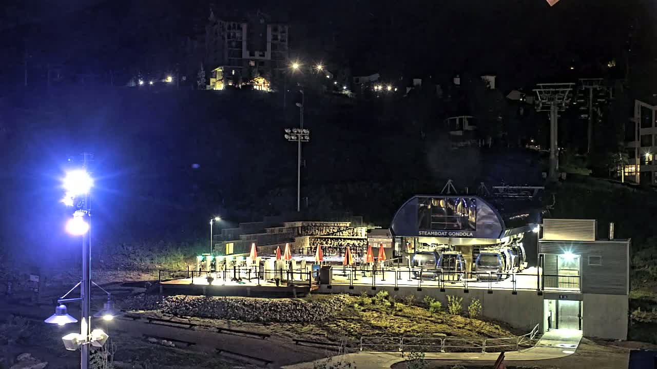 A gondola station with several cars illuminated at night, situated on a hillside with buildings and ski lifts visible in the background under dark skies.