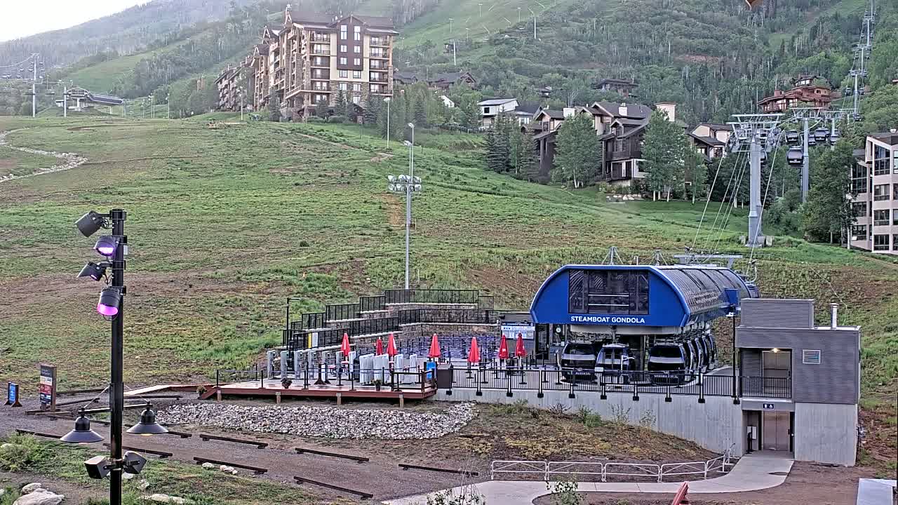 A gondola station sits at the base of a grassy hill dotted with buildings, under a cloudy sky.