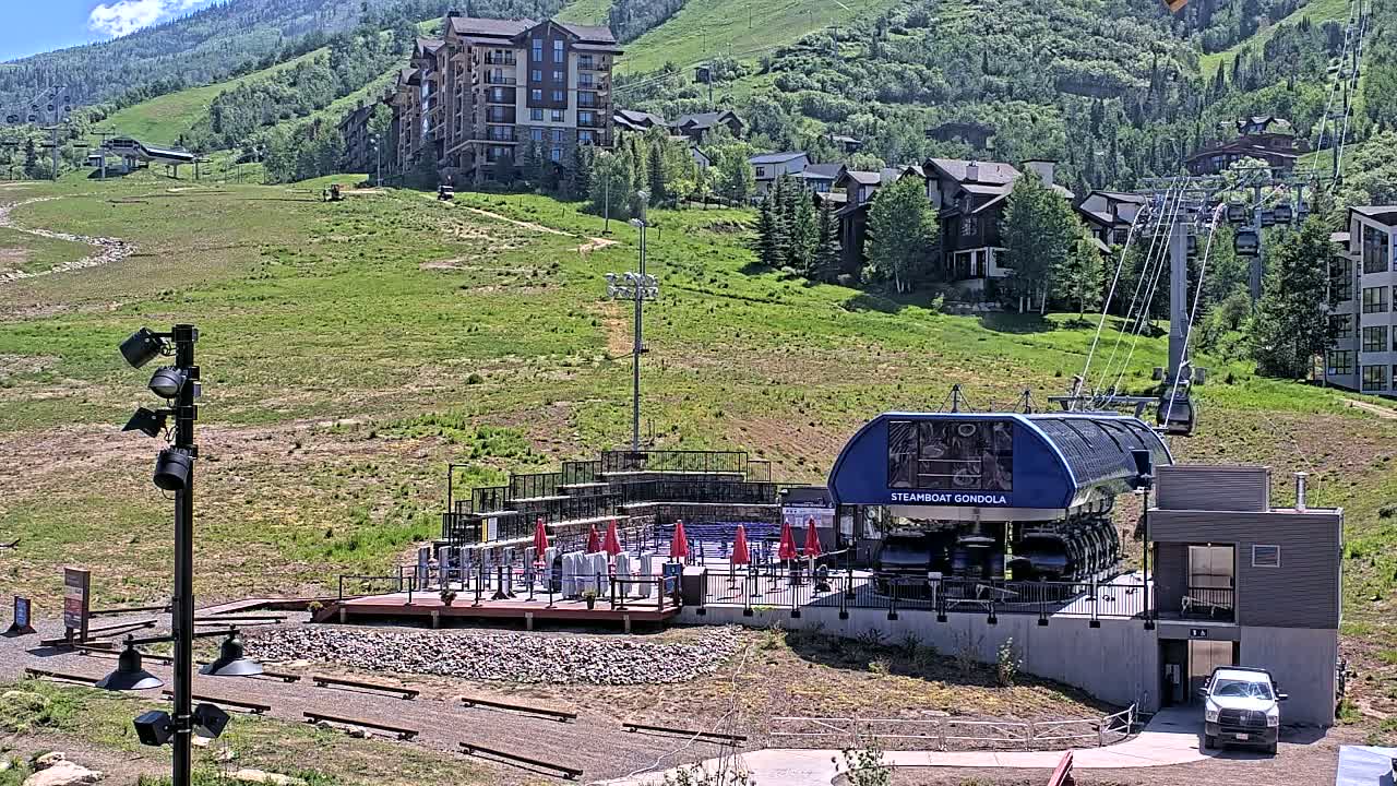 A gondola station sits at the base of a grassy mountainside with buildings and ski lifts in the background on a sunny day.