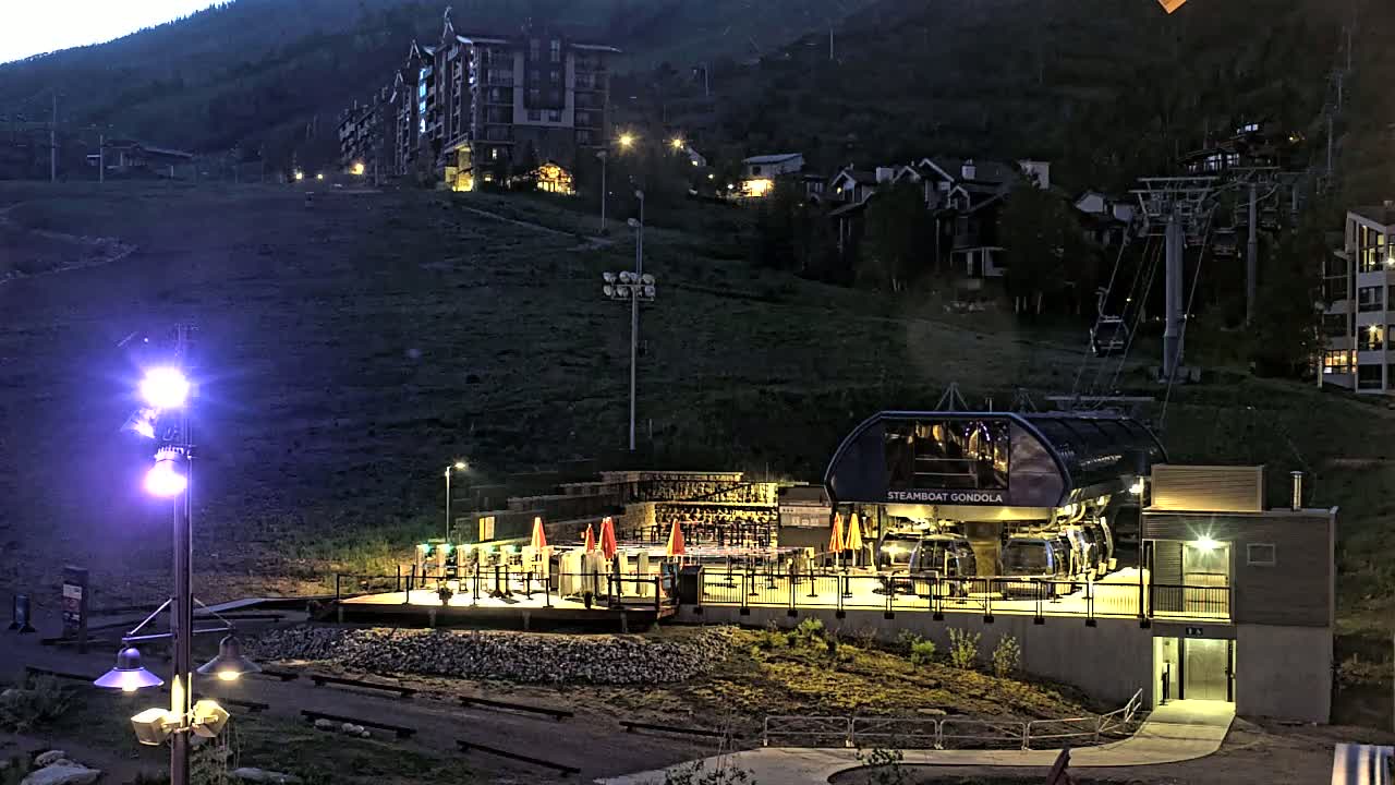 A gondola station at night, illuminated by artificial lights, sits on a hillside overlooking a town nestled in a mountain valley under a dark sky.