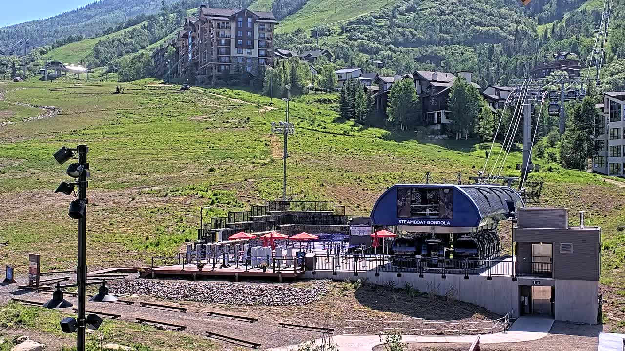 A gondola station sits on a grassy hillside, under a bright, sunny sky, with a mountain and residential buildings visible in the background.