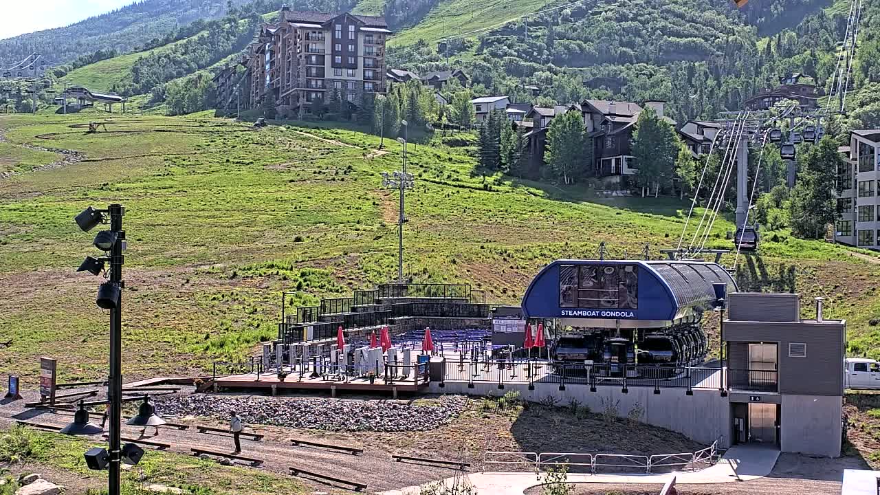 A gondola station sits at the base of a grassy mountainside dotted with buildings, under a sunny sky.