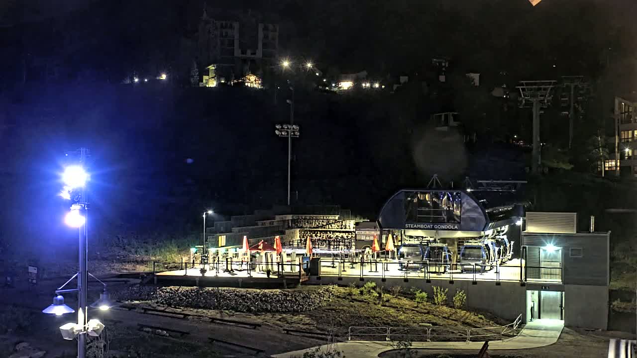At night, a gondola station with several gondolas sits at the base of a hill with buildings and lights visible on the hillside.