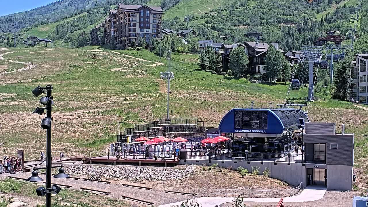 A gondola station sits on a grassy hillside under hazy, sunlit conditions, with a chairlift visible in the background.