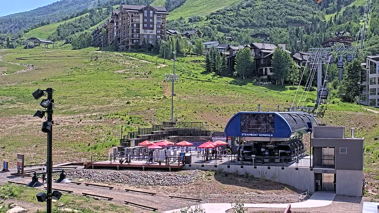 A gondola station sits below a hillside of buildings and green grass under a sunny sky.