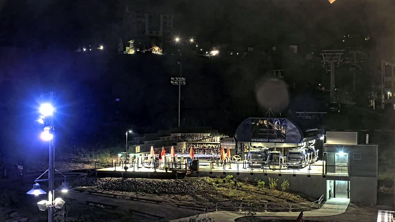 A gondola station, illuminated at night, sits at the base of a hill with lights visible on the hillside and a ski lift in the background.