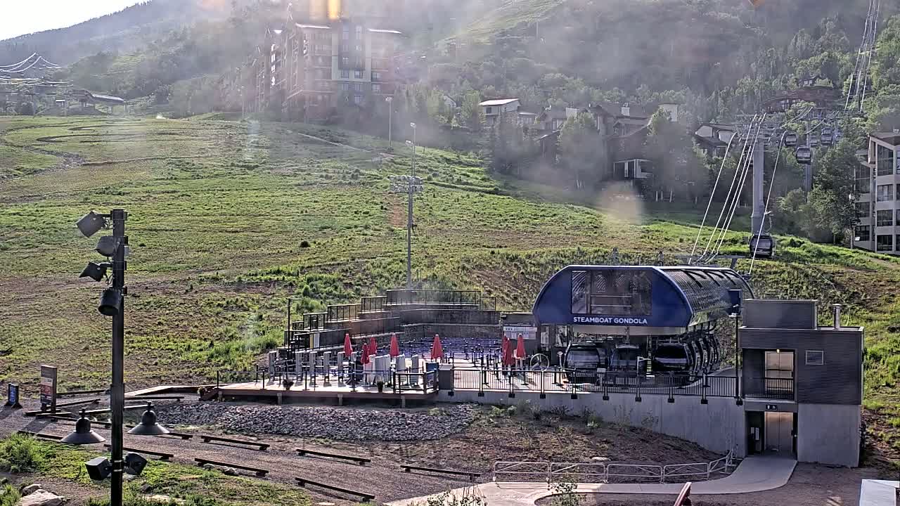 A gondola station sits at the base of a grassy mountainside with buildings and a cable car system visible in the hazy, sunny weather.