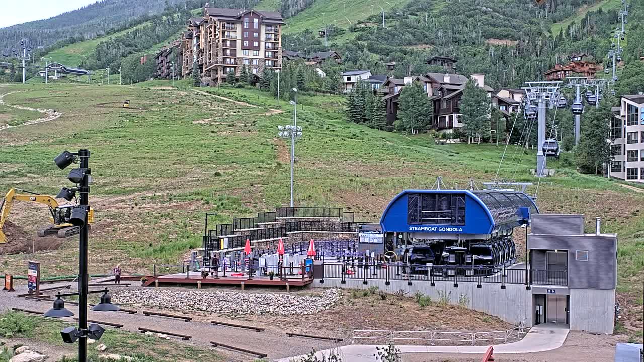 A gondola station sits on a grassy hillside below a cluster of buildings and ski lifts, under a mostly clear sky.