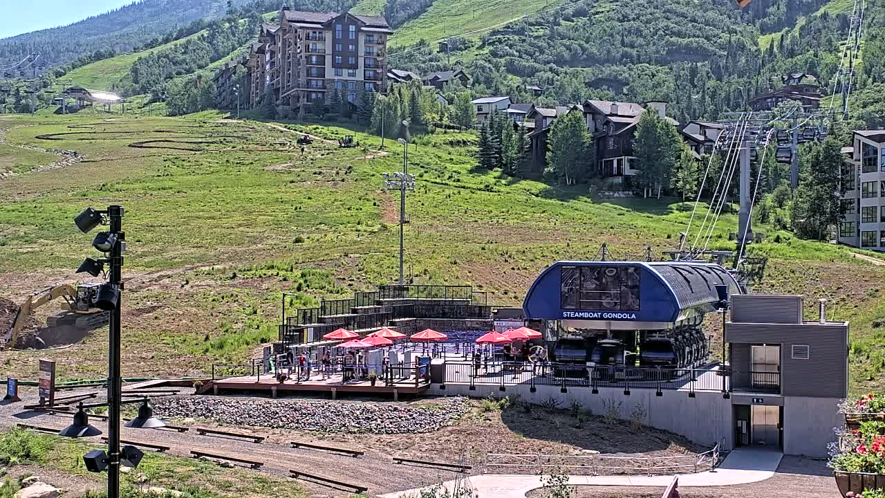 A gondola station with red umbrellas and outdoor seating sits on a grassy hillside below a cluster of buildings on a sunny day.