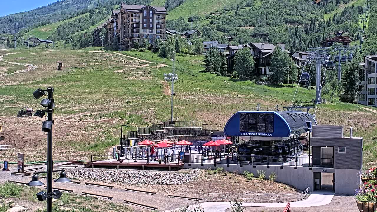 A gondola station sits at the base of a grassy mountainside on a sunny day, with a chairlift ascending the hill behind it.