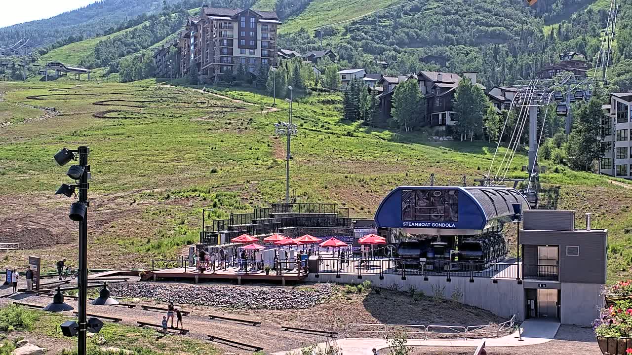 A gondola station sits at the base of a grassy mountainside with several buildings and a chairlift in the background on a sunny day.
