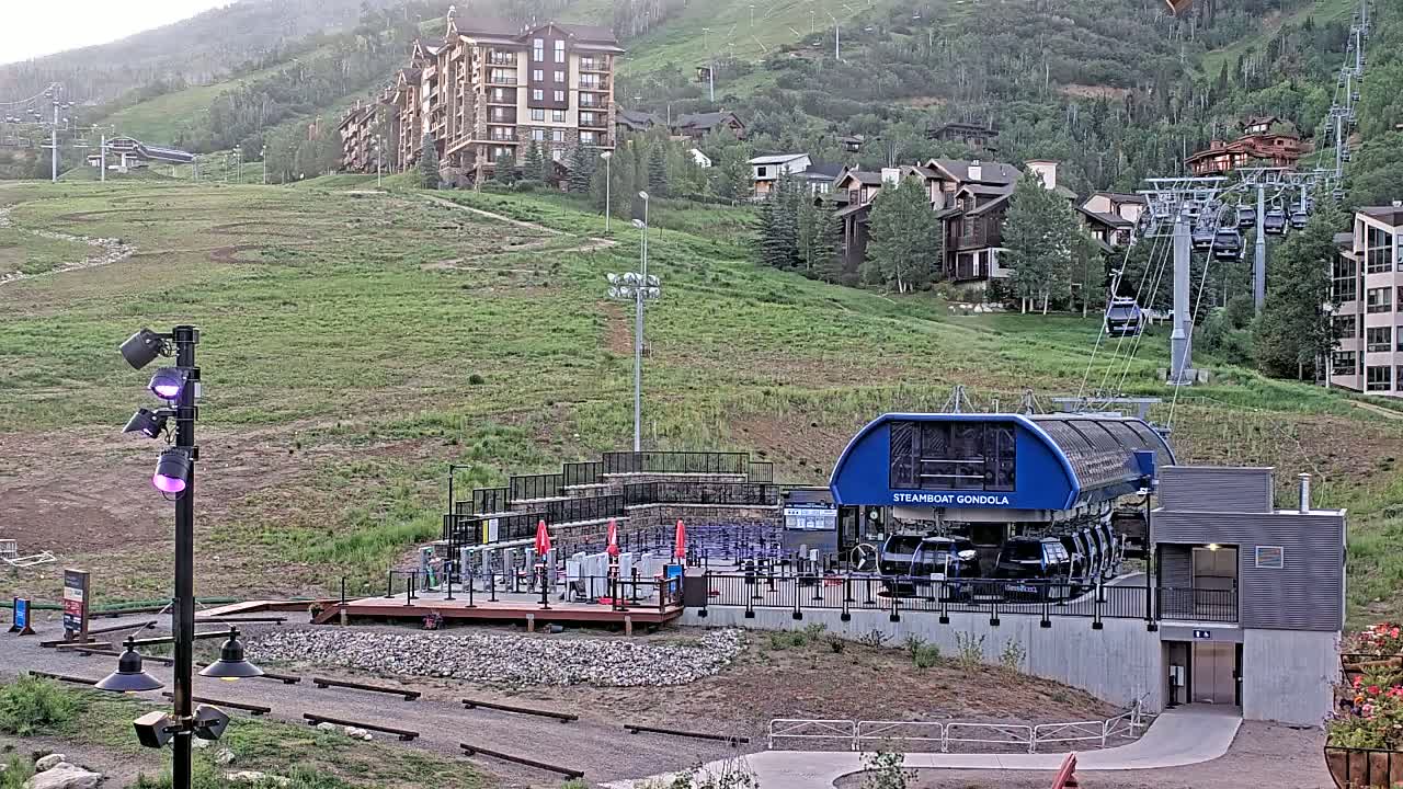A gondola station sits on a grassy hillside with a ski lift and buildings in the background, under a partly cloudy sky.