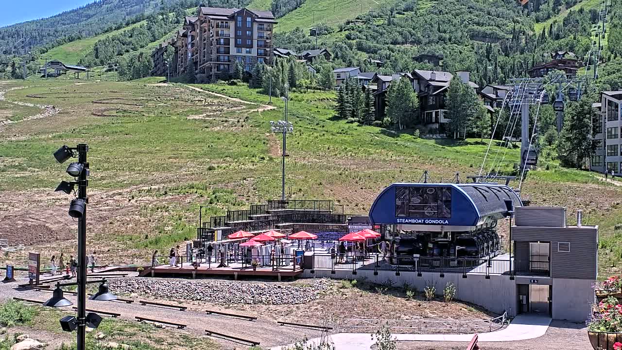 A gondola station with a blue gondola car sits below a mountainside of homes and green hills under a sunny sky.