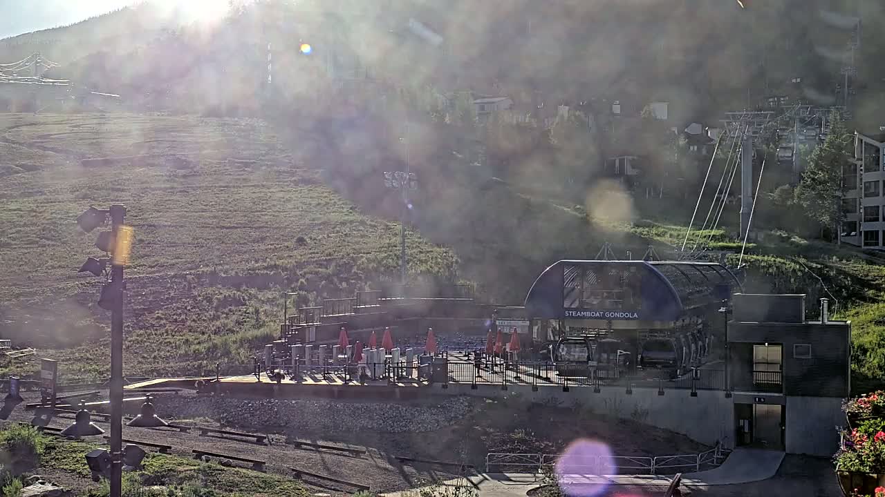 A gondola station sits at the base of a sun-drenched, hazy mountain, with a chairlift visible further uphill.