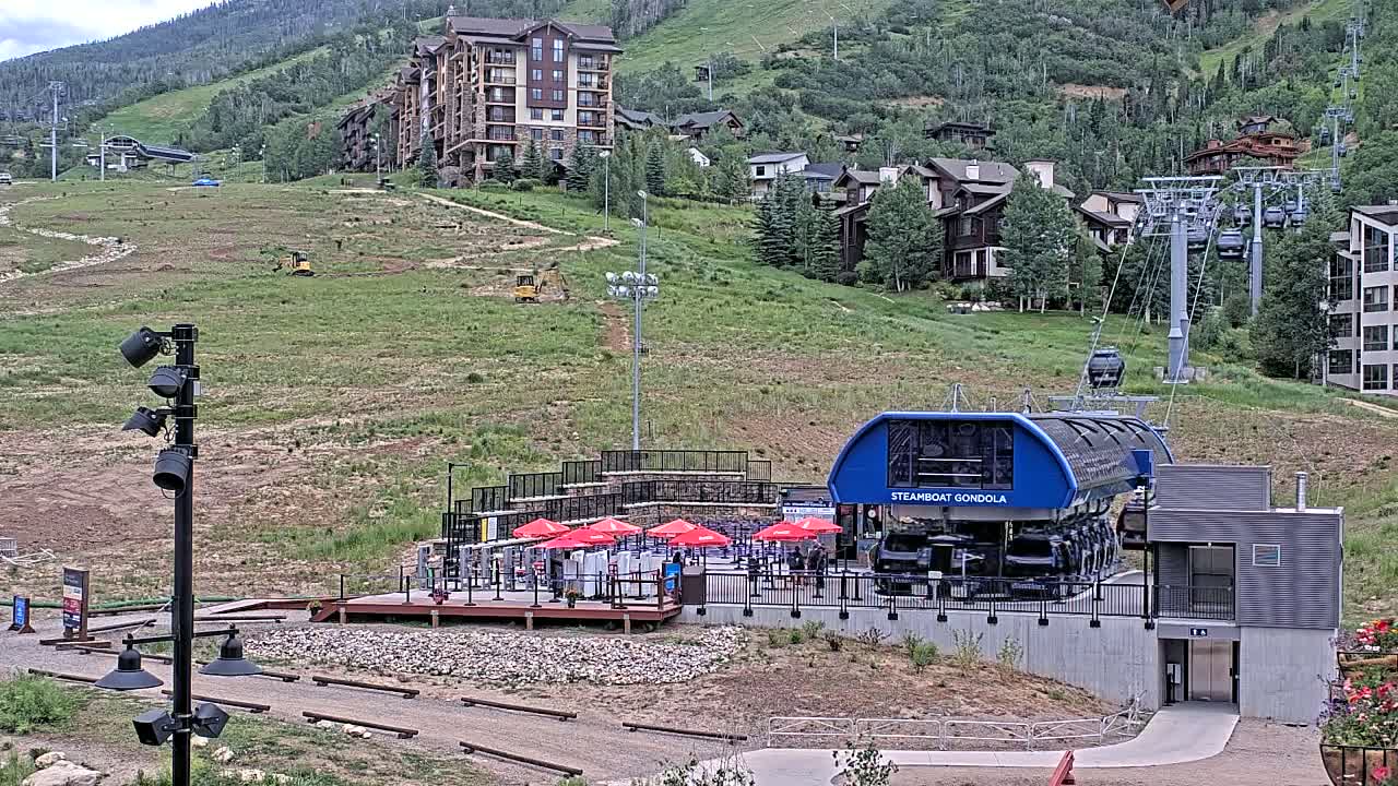 A gondola station with red umbrellas sits on a grassy hillside below a mountain resort, under partly cloudy skies.