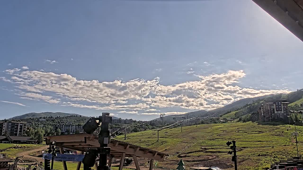 A mostly sunny sky with scattered clouds overlooks a grassy hillside with a ski lift and buildings in the distance.