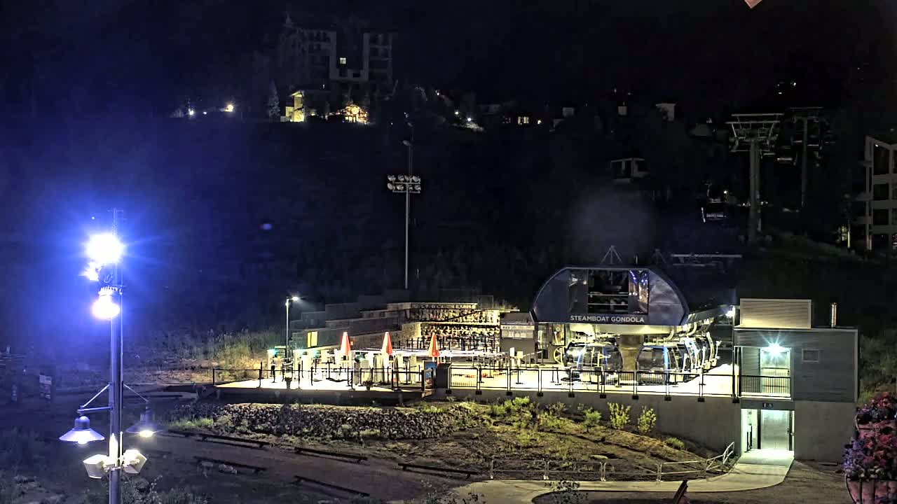 At night, a gondola station with its cars illuminated sits on a hillside below buildings, under a clear, dark sky.