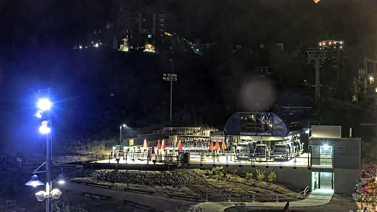 At night, a gondola station with its cars illuminated sits on a hillside below buildings, under a clear, dark sky.