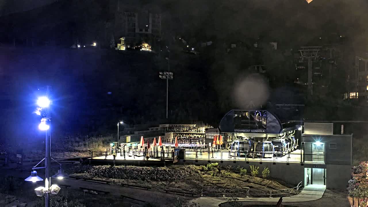 At night, a gondola station with several passenger cars is illuminated, situated on a hillside with buildings visible in the background.