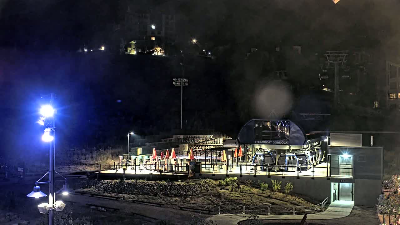 At night, a gondola station with several passenger cars is illuminated, situated on a hillside with buildings visible in the background.