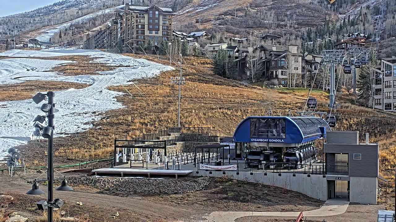 At night, a gondola station with several passenger cars is illuminated, situated on a hillside with buildings visible in the background.