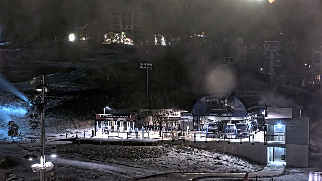 A brightly lit gondola station stands amidst snow-covered slopes at a ski resort at night, with snow guns actively producing fresh powder under foggy conditions, and distant illuminated buildings dotting the hills.