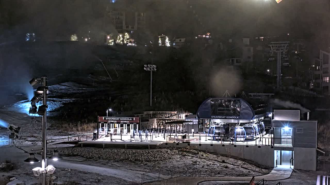 A brightly lit gondola station stands at the base of a ski slope where snowmaking machines are actively producing snow under hazy night conditions, with illuminated resort buildings visible on the dark hillside in the background.