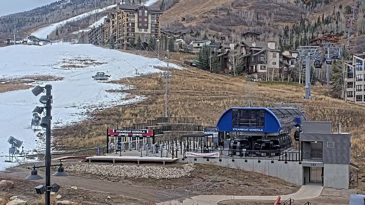 Under an overcast sky, a ski resort scene reveals a large blue gondola station and a snow-covered ski run with a visible snow groomer, set against dry, brown hillsides dotted with various buildings and ski lifts.