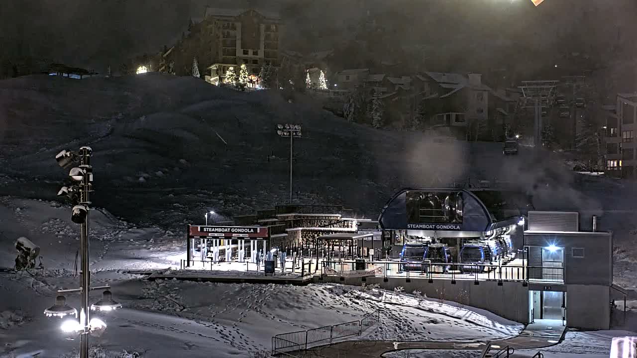 A snowy mountain ski resort gondola station is brightly lit at night, with multiple buildings and ski lifts visible on the slopes through a light mist or fog.