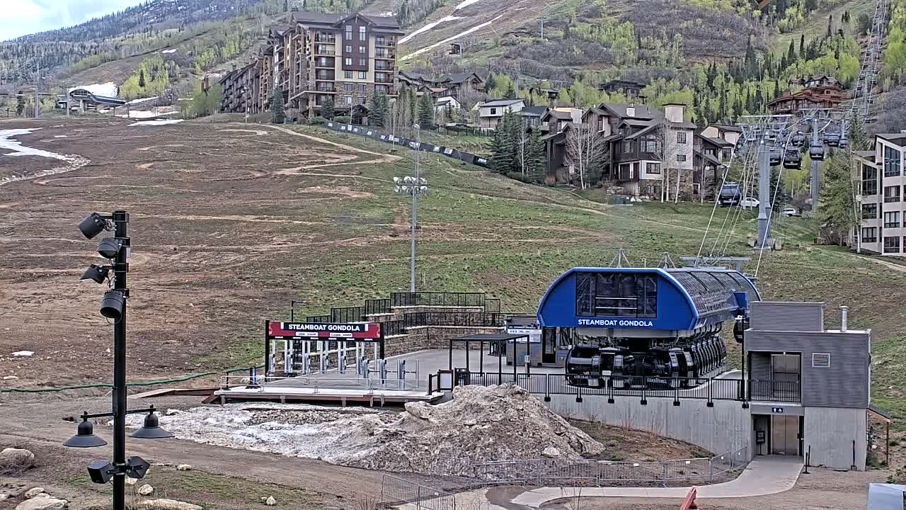 A snowy mountain ski resort gondola station is brightly lit at night, with multiple buildings and ski lifts visible on the slopes through a light mist or fog.