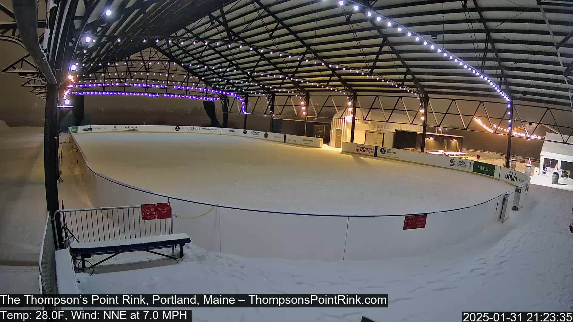 A snow-covered outdoor ice rink is illuminated at night under a metal structure with string lights, during a snowfall with a temperature of 28°F and a north-northeast wind.