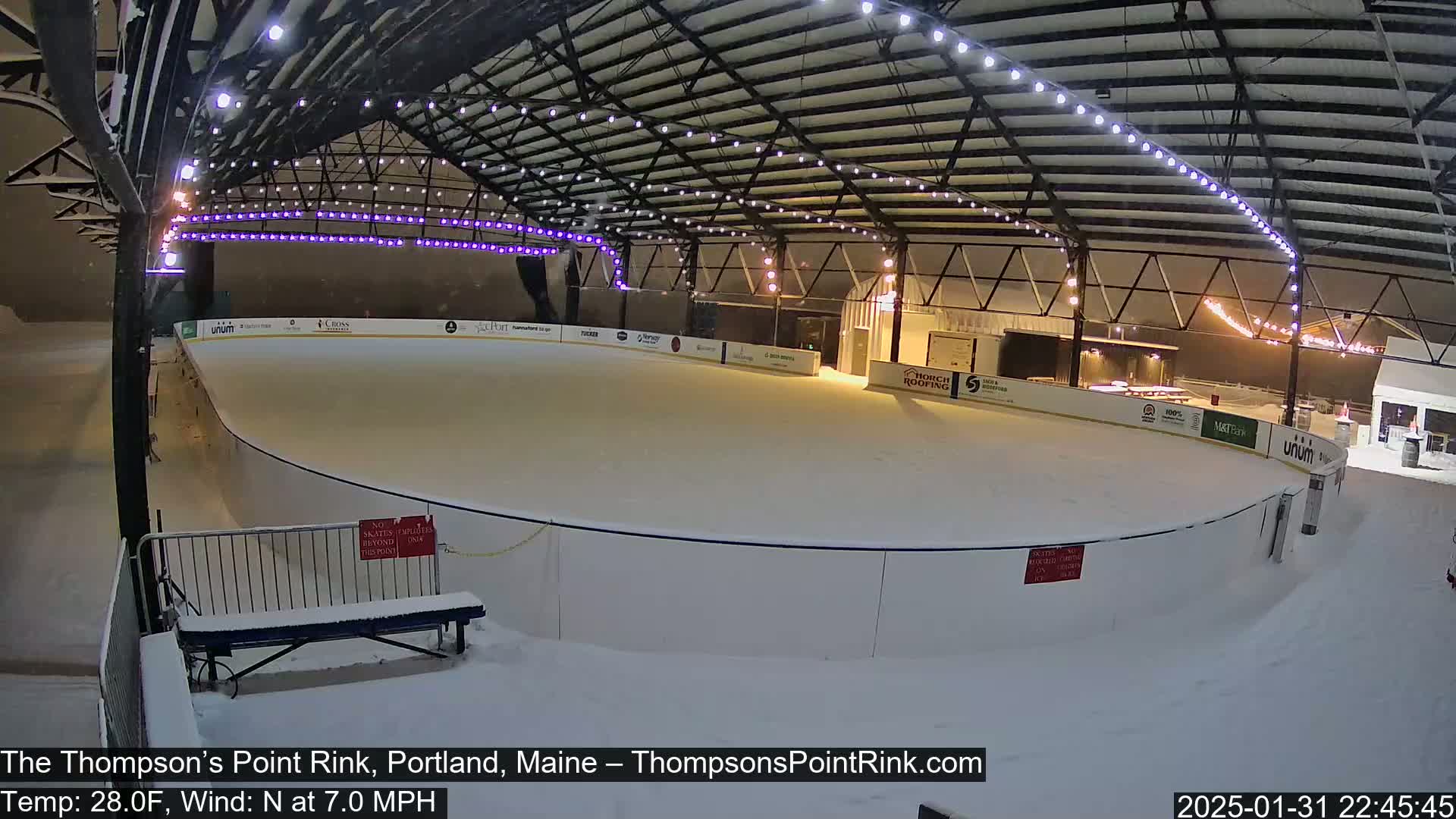 A snow-covered outdoor ice rink is illuminated at night under a metal structure with string lights, during a snowfall with a north wind.
