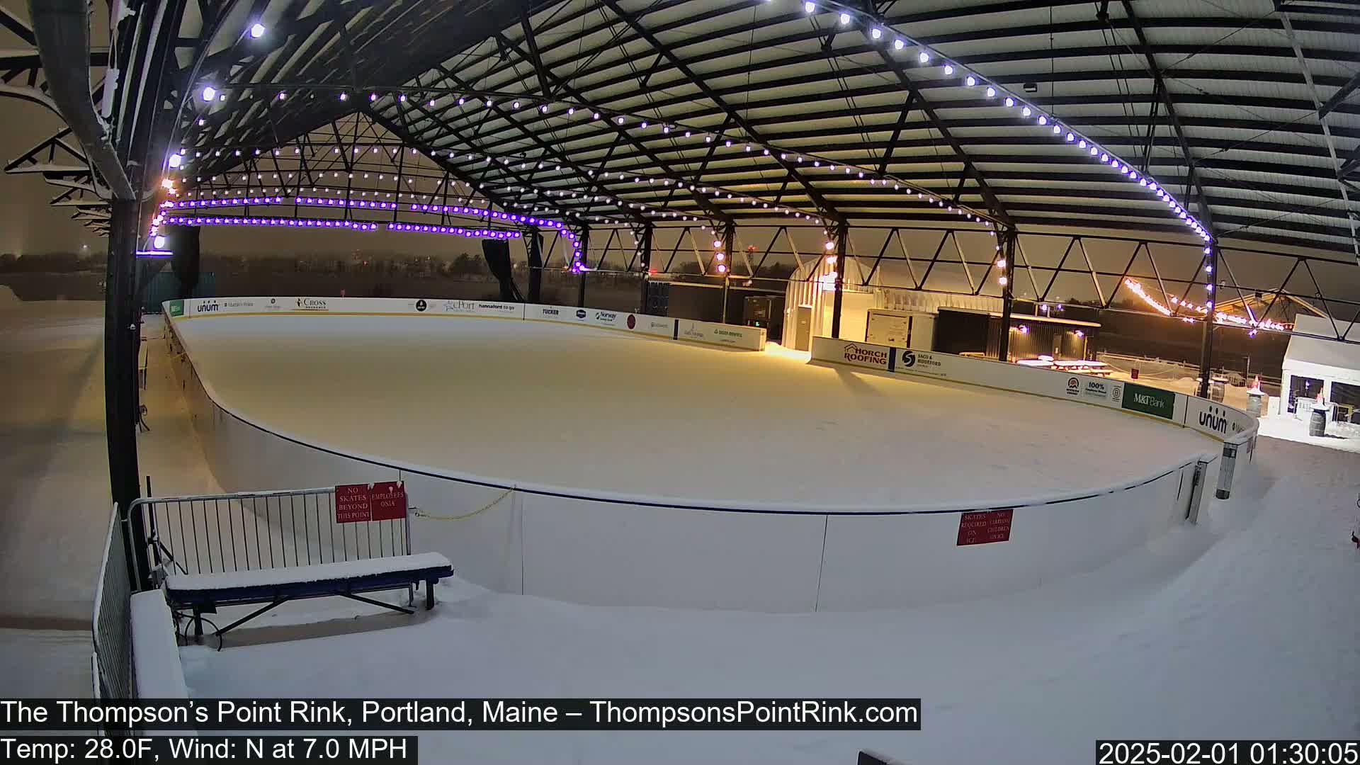 A snow-covered outdoor ice rink is illuminated at night under a metal-framed structure with string lights.