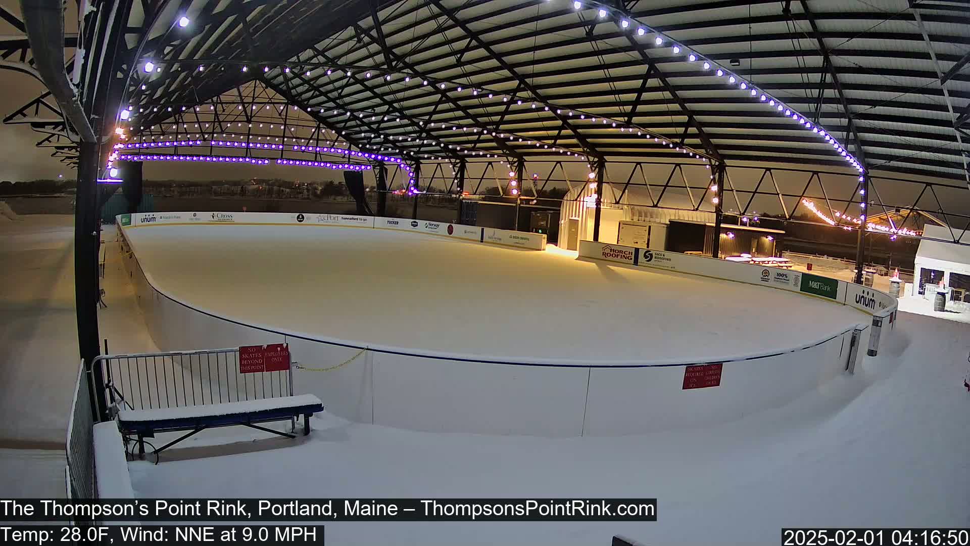 A snow-covered outdoor ice rink is illuminated at night under a metal structure with string lights.