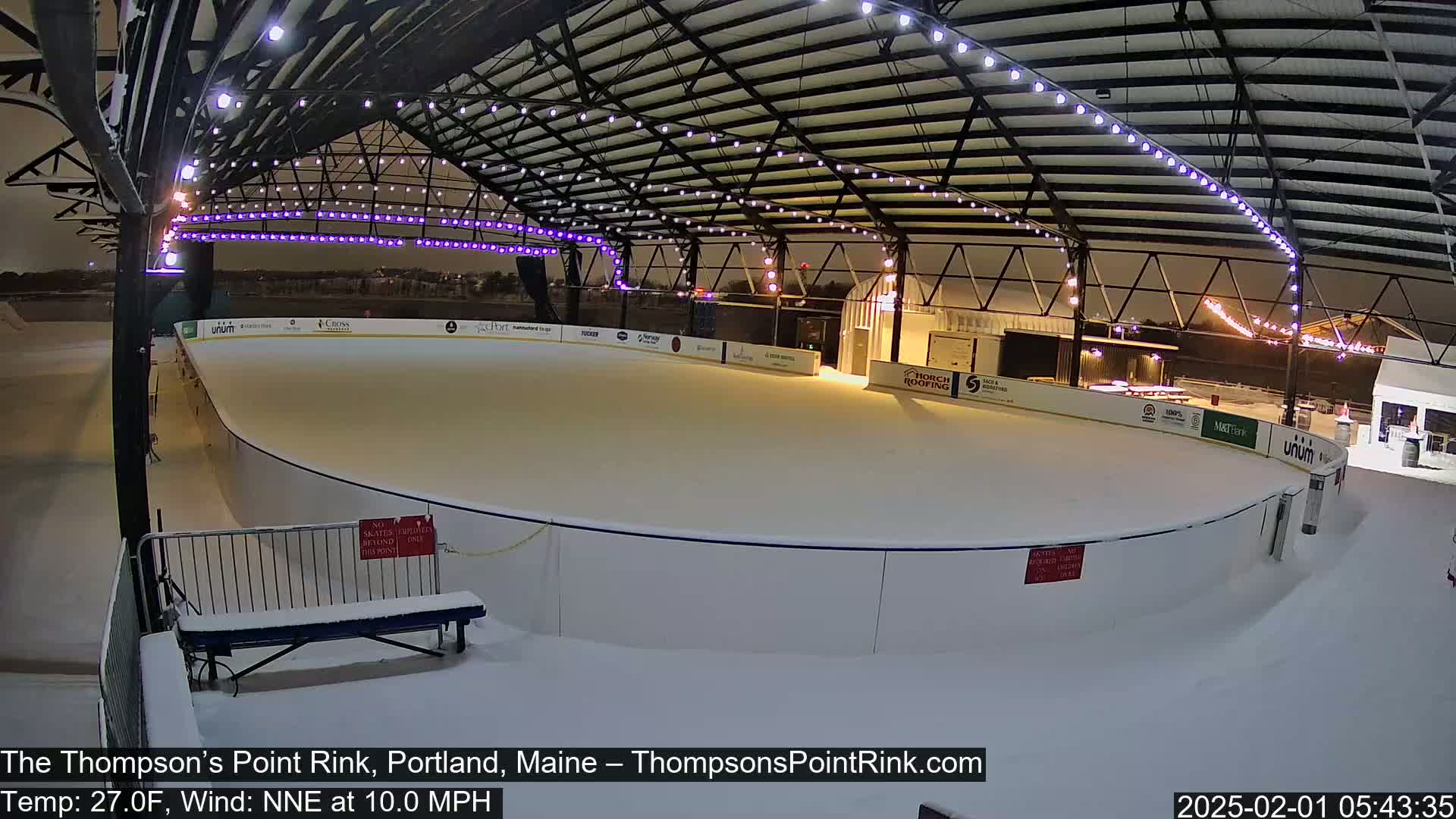 A snow-covered outdoor ice rink is illuminated at night under a metal structure with string lights.