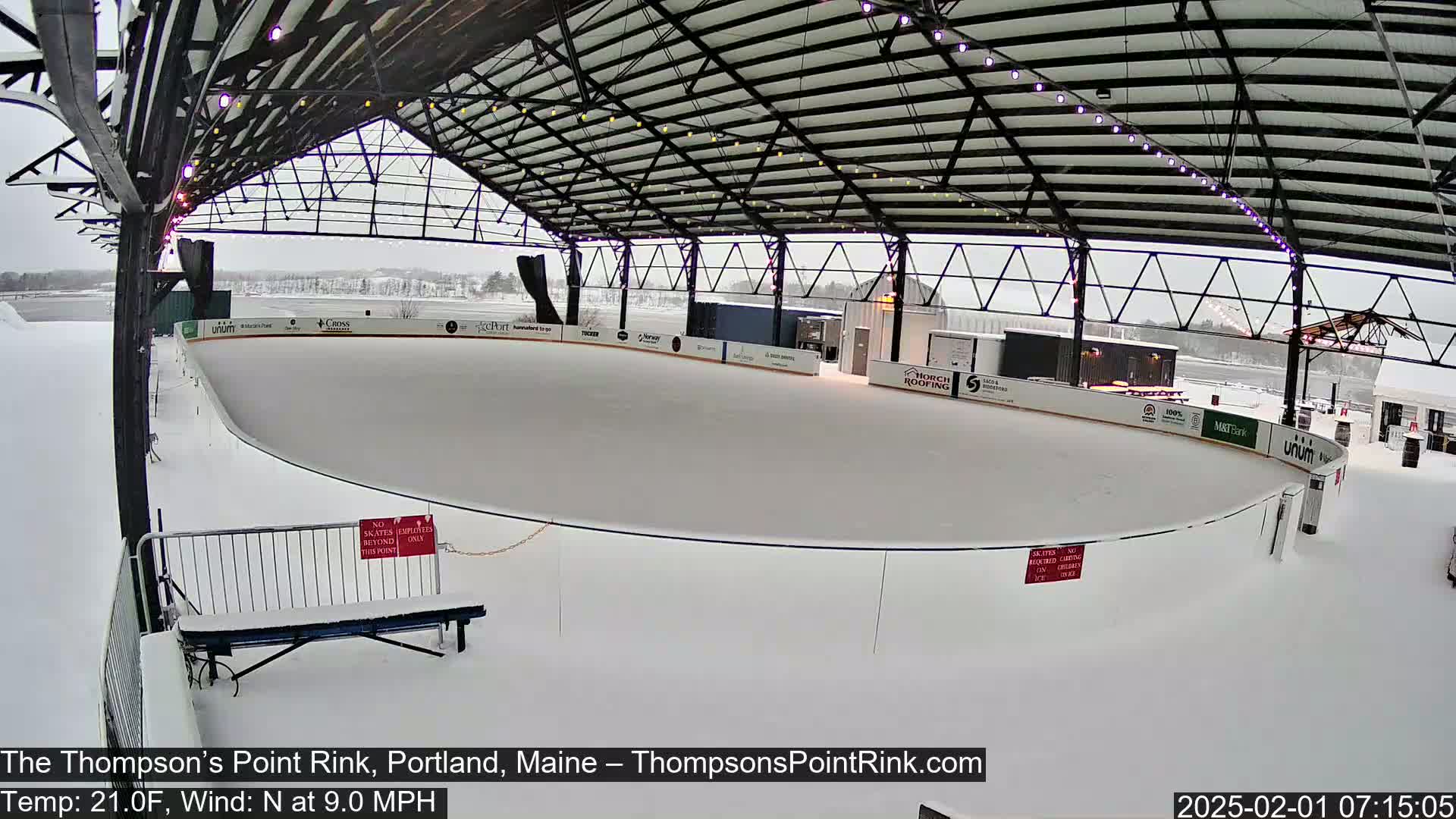 A snow-covered outdoor ice rink sits under a metal structure during a light snowfall.