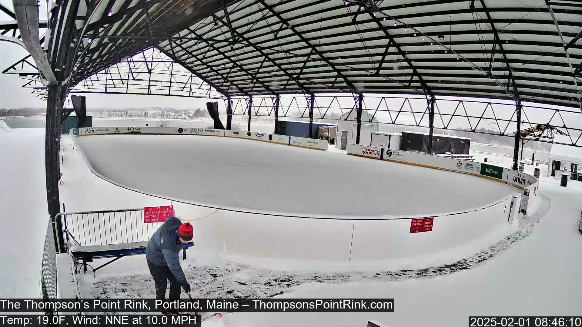 A person is clearing snow from around an outdoor ice rink under a metal roof during a light snowfall.