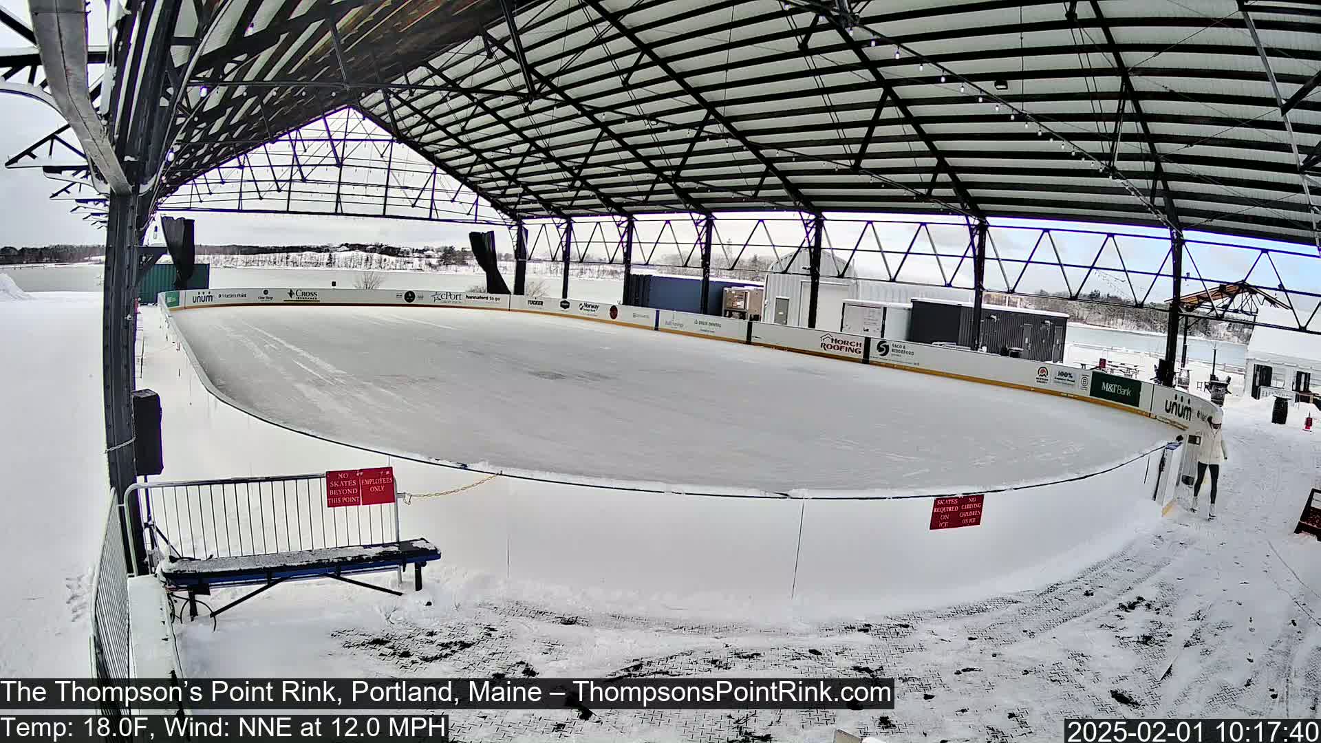 An outdoor ice rink, covered by a metal structure, is surrounded by snow, under a partly cloudy sky.