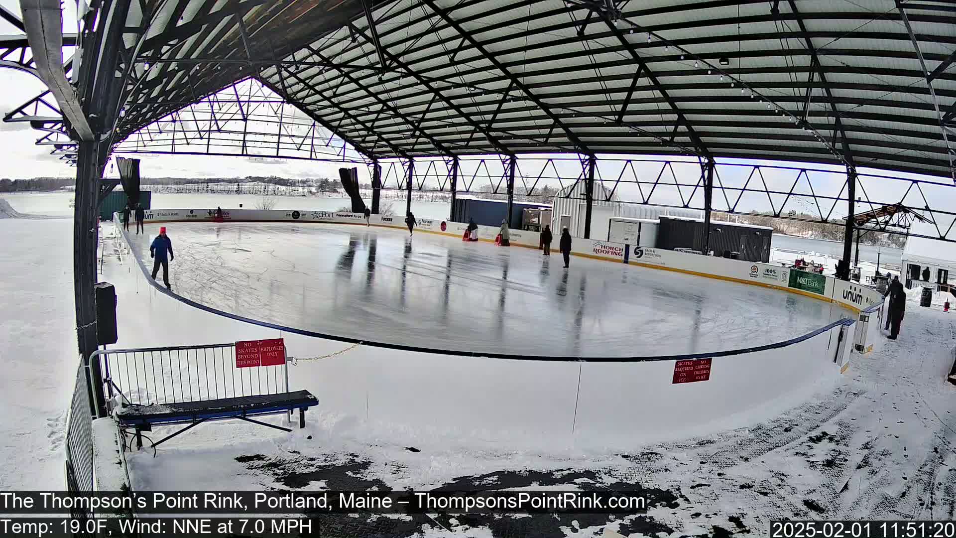An outdoor ice rink, partially covered by a metal structure, is surrounded by snow and has several people ice skating on it.