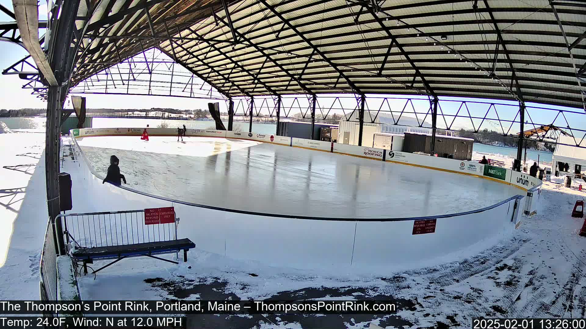 A partially covered outdoor ice rink, surrounded by snow, under a sunny sky.
