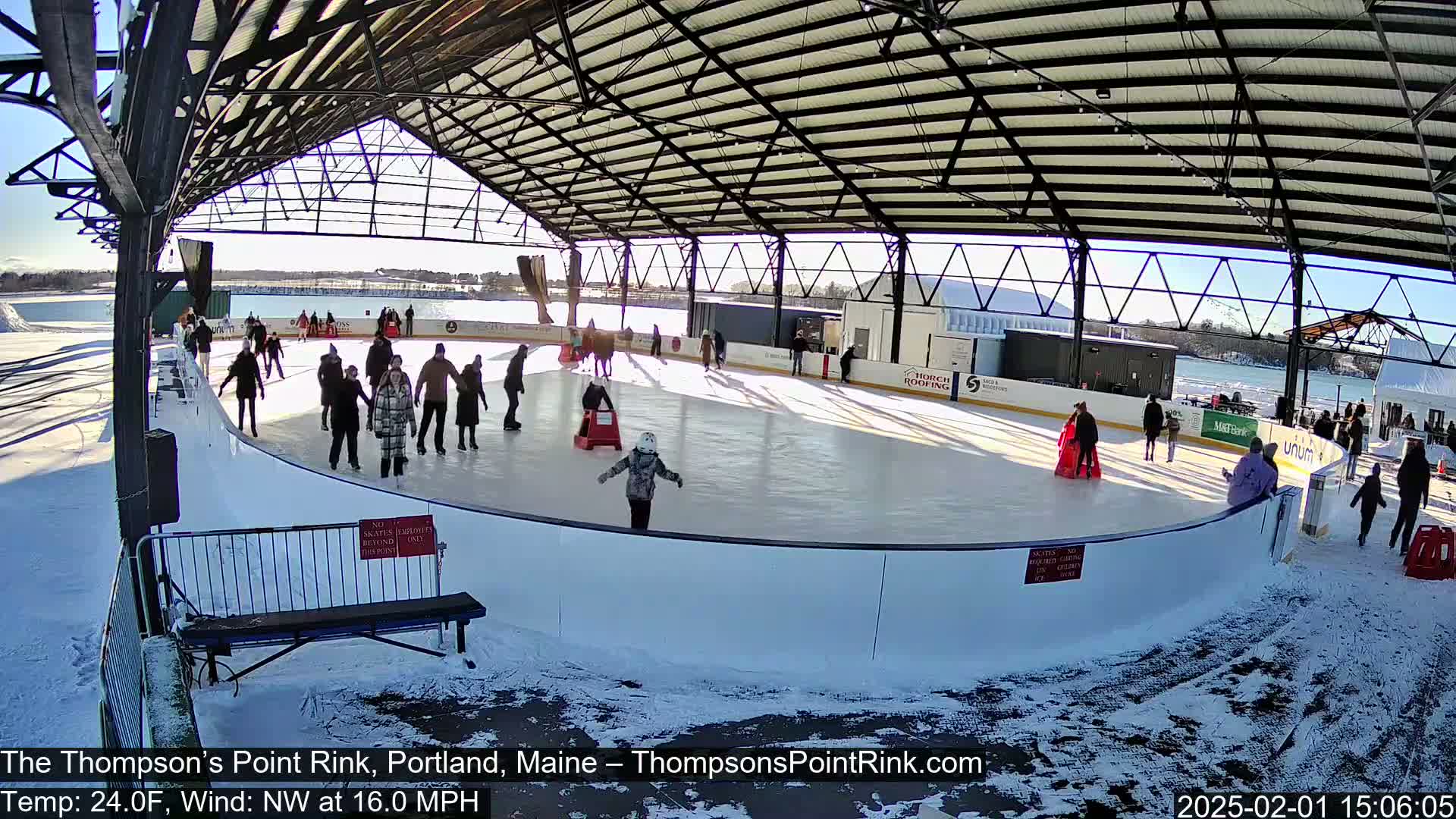 Under a sunny sky, several people ice skate on an outdoor rink partially enclosed by a metal structure, with snow on the ground around the rink.