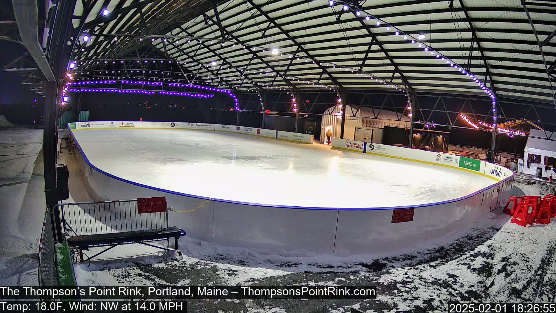 An outdoor ice rink, lit with purple string lights, is covered in snow with a 14 mph northwest wind and 18°F temperature.