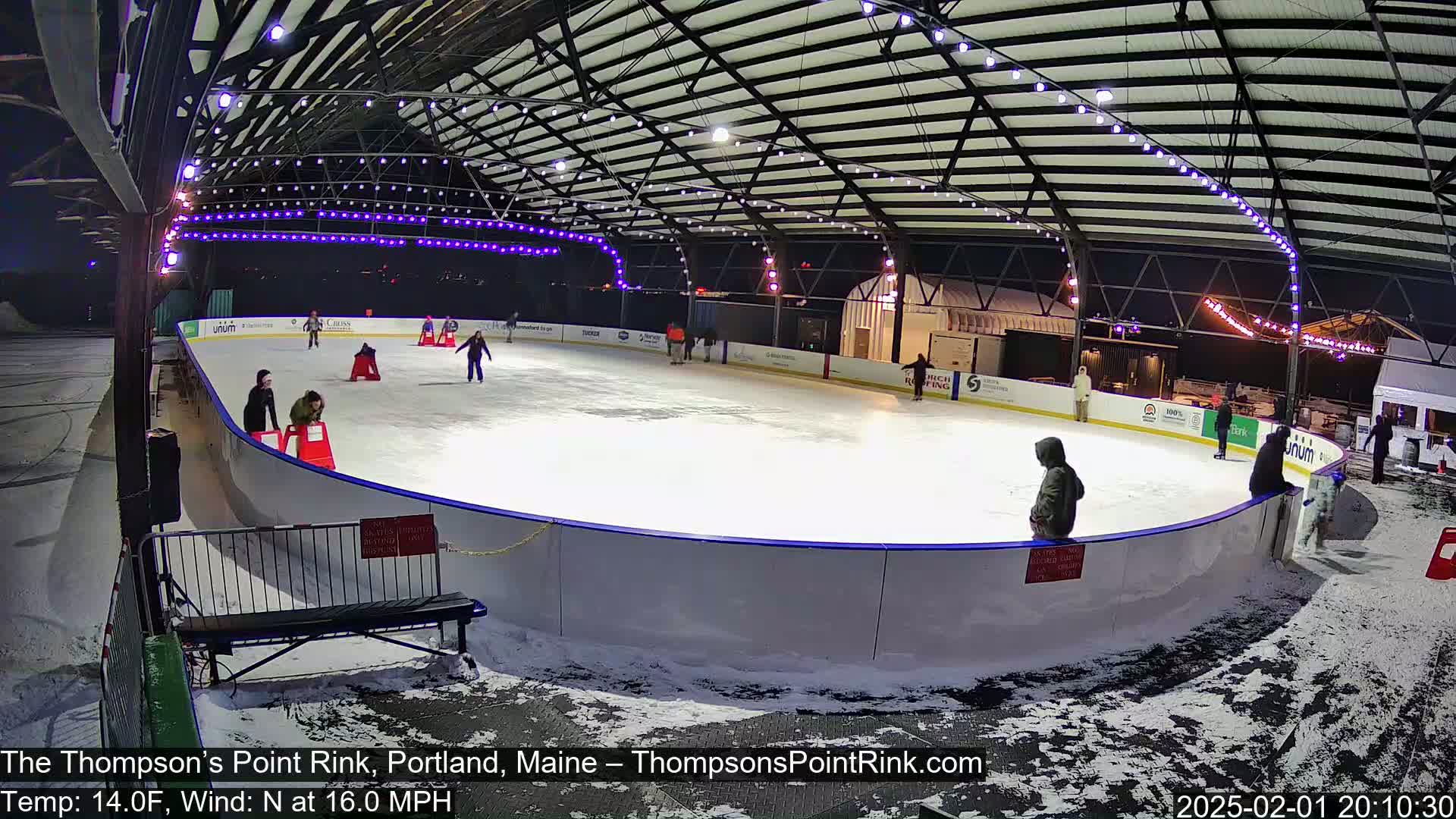 An outdoor ice skating rink, illuminated at night under a metal structure, with several people ice skating and some standing around the rink's perimeter in snowy conditions.