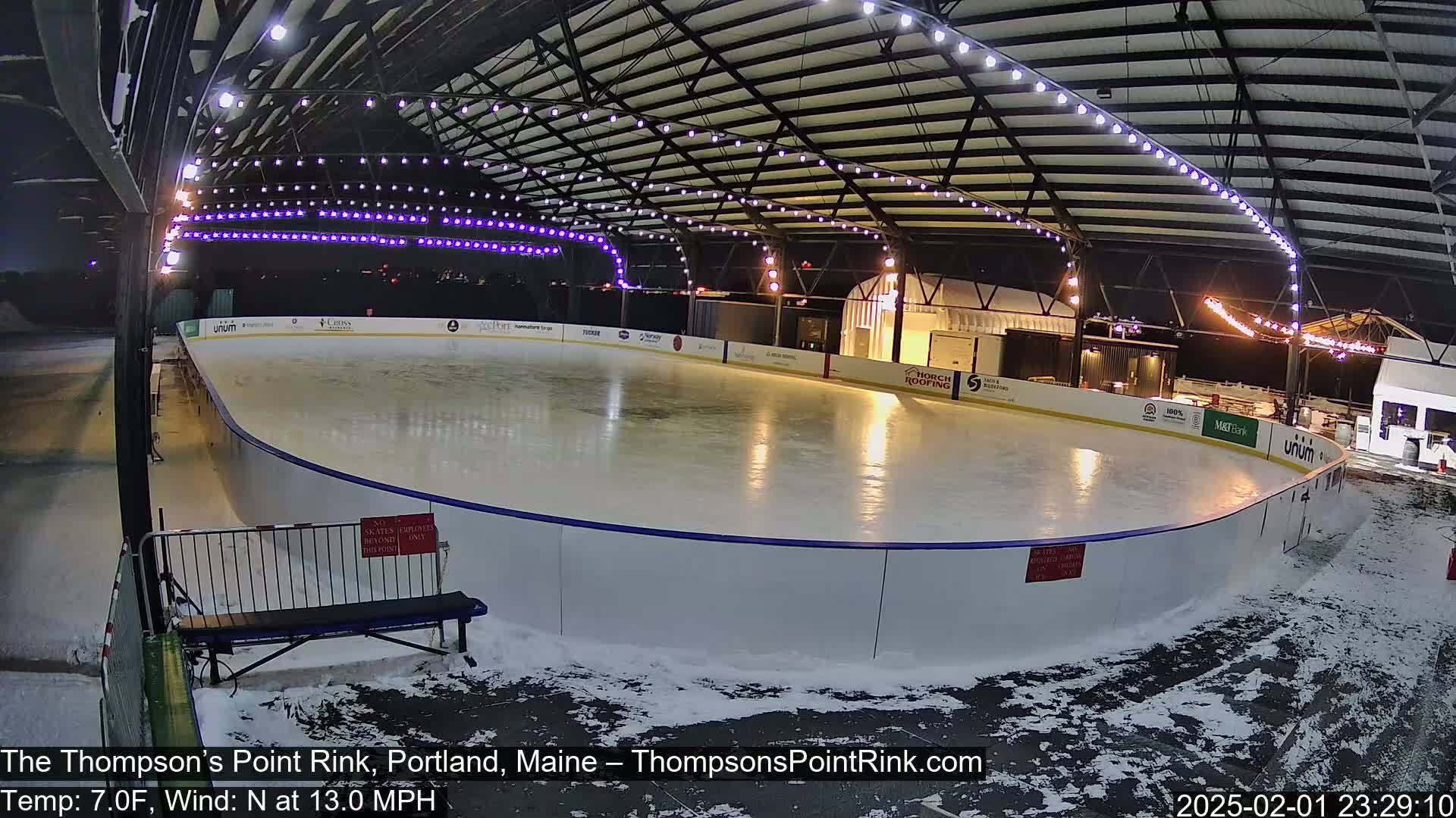 An outdoor ice rink, lit at night under a metal structure, is partially covered in snow with a slight north wind.