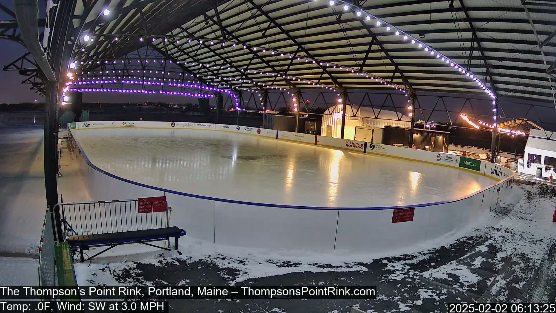An outdoor ice rink, under a metal structure with string lights, is partially covered in snow at night.