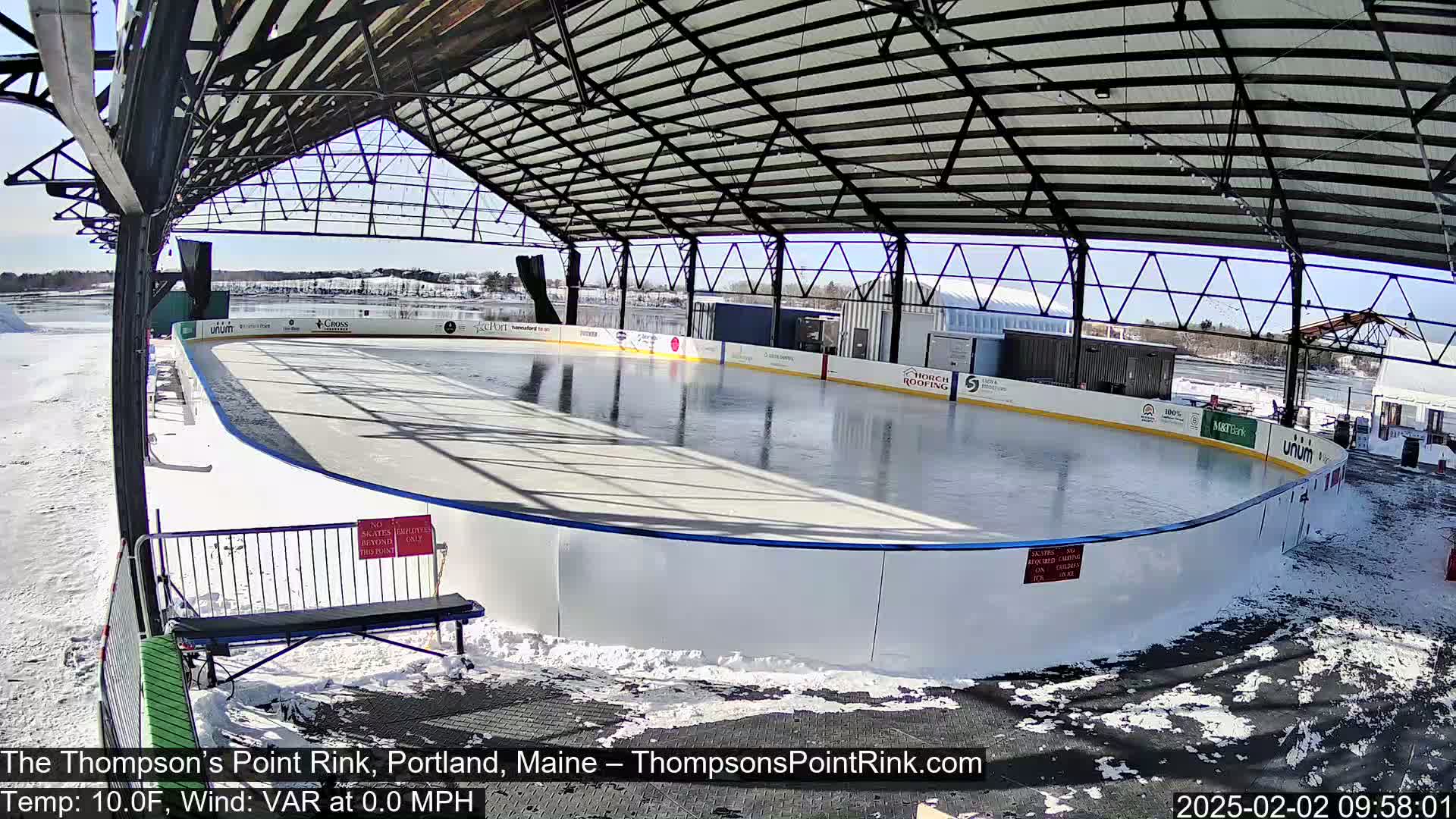 A sunny day reveals an outdoor ice rink enclosed by a metal structure, surrounded by snow.