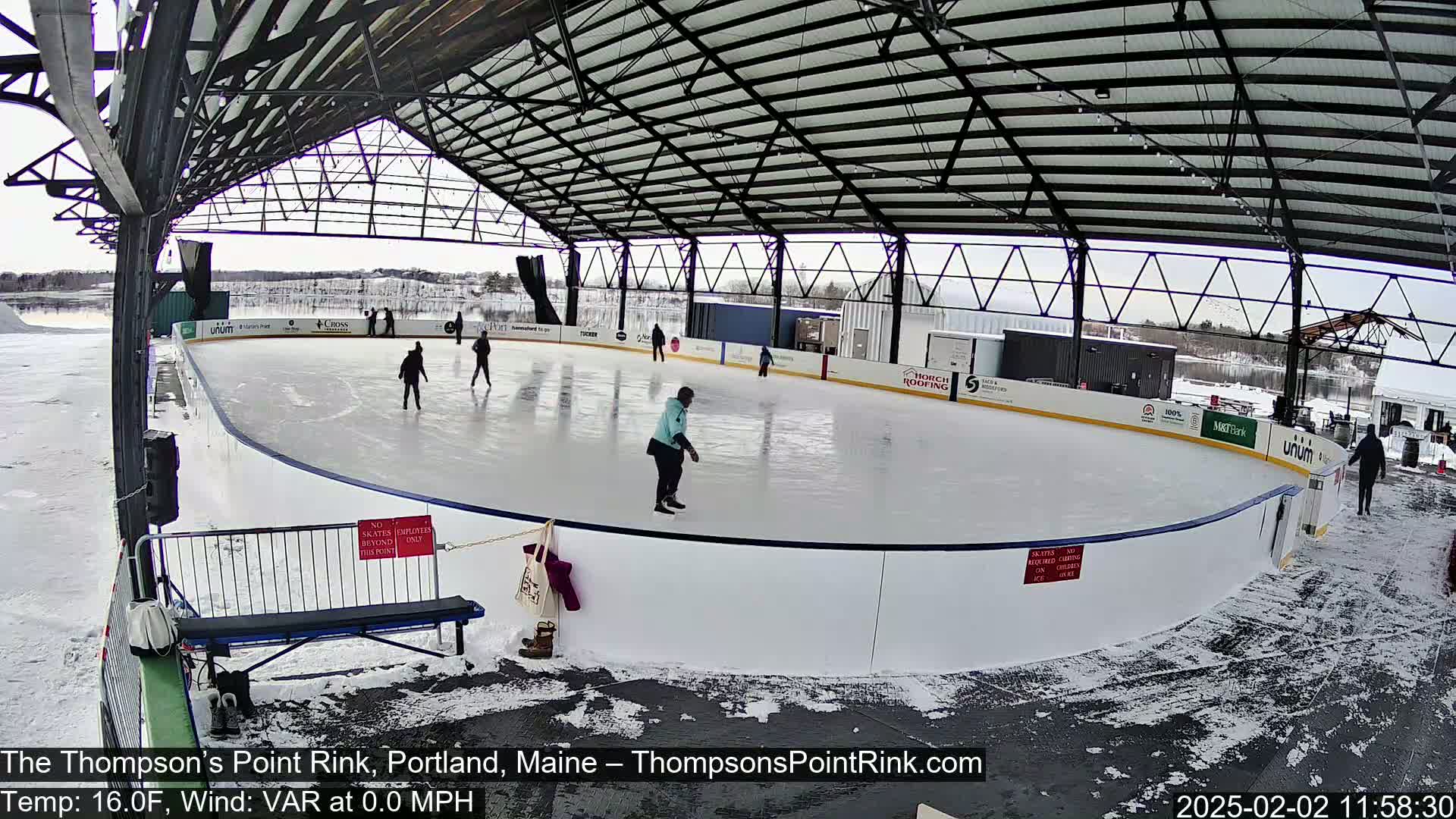 A covered outdoor ice rink with several people ice skating on a partly snowy day.