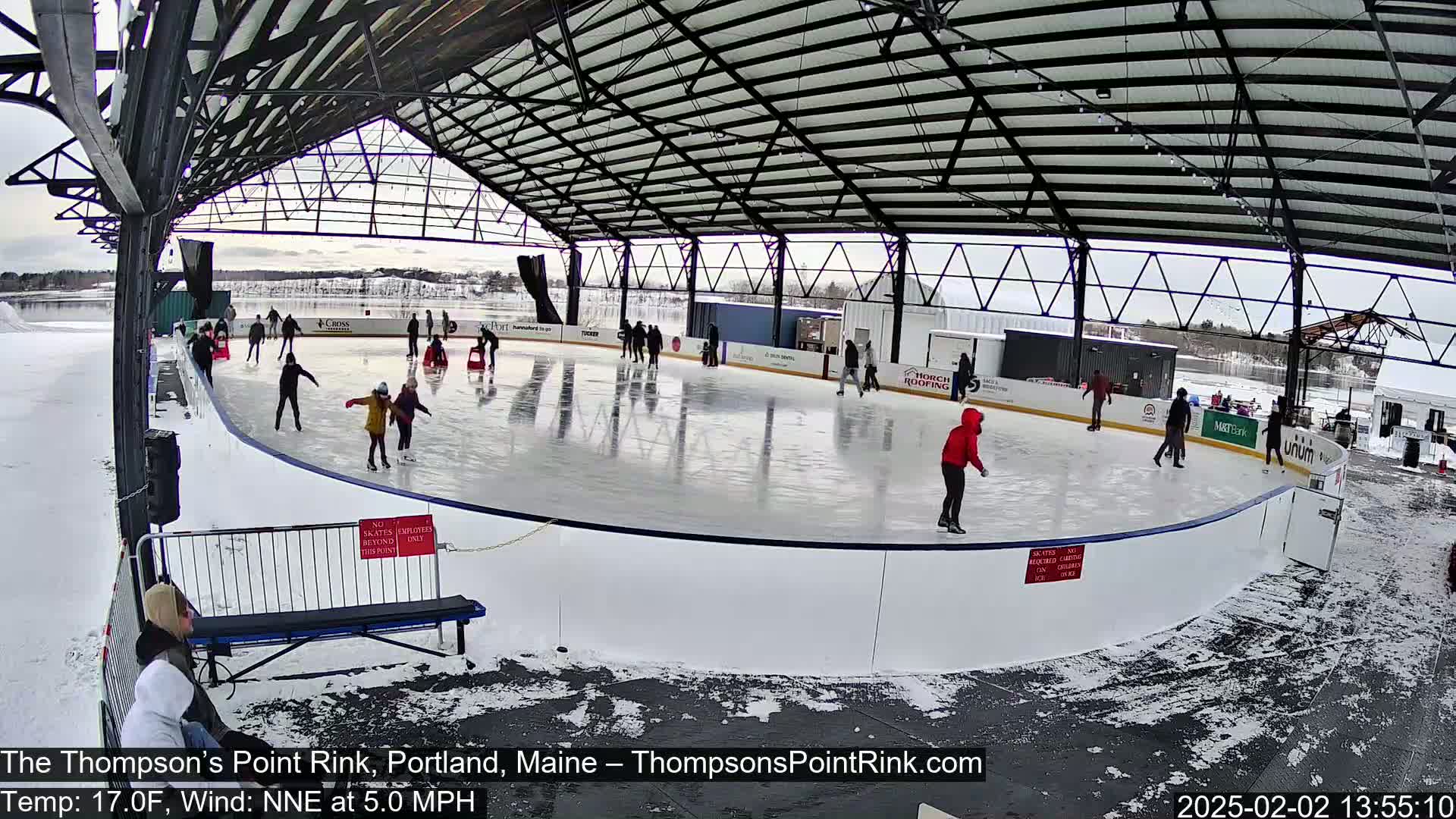 An outdoor ice skating rink, partially covered by a metal structure, is populated by several skaters on a partly sunny, cold day.