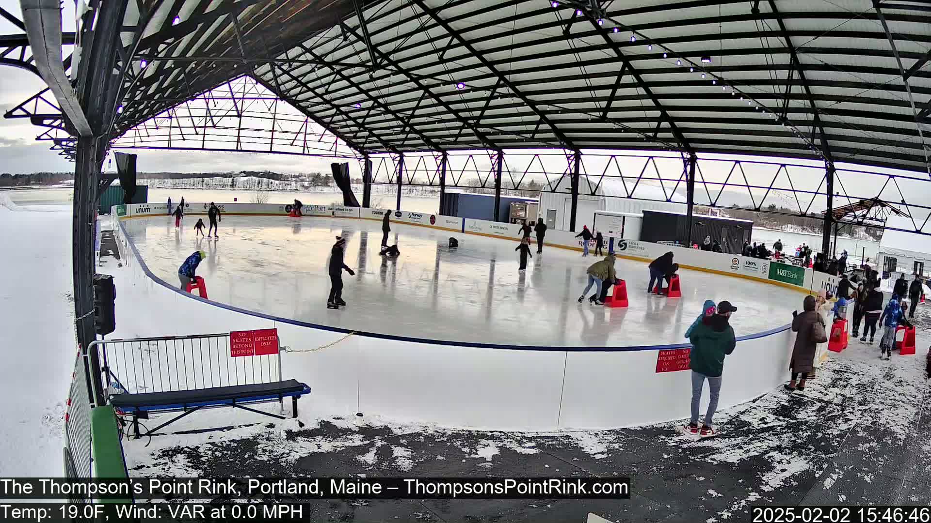 Many people ice skate on an outdoor rink under a metal roof on a partly cloudy day with a temperature of 19°F and no wind.