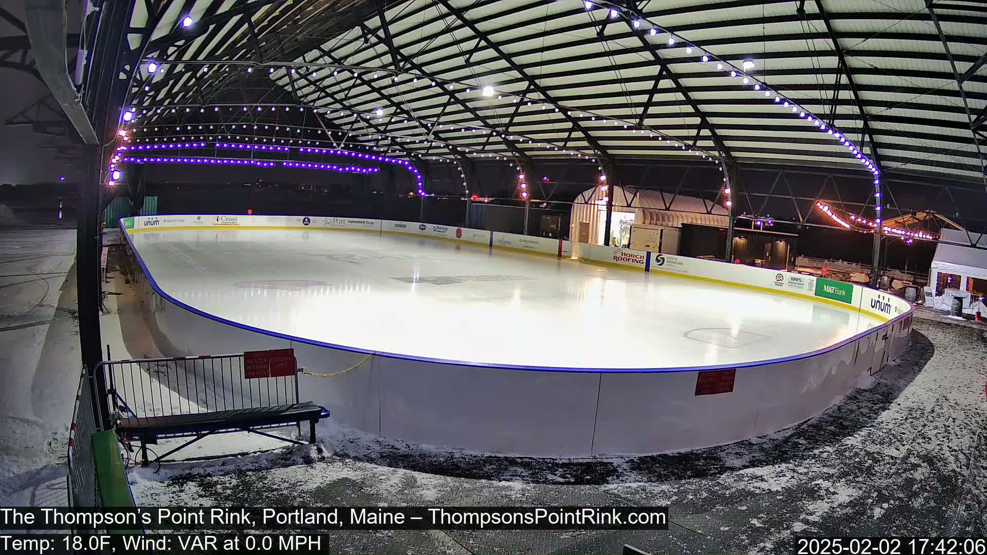 An outdoor ice rink, illuminated at night under a metal structure, with light snow on the ground.